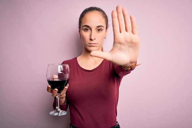 Free photo young beautiful woman drinking glass with red wine over isolated pink background with open hand doing stop sign with serious and confident expression defense gesture