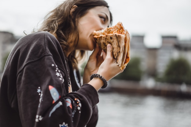 Giovane bella donna che mangia una fetta di pizza sulla via della città