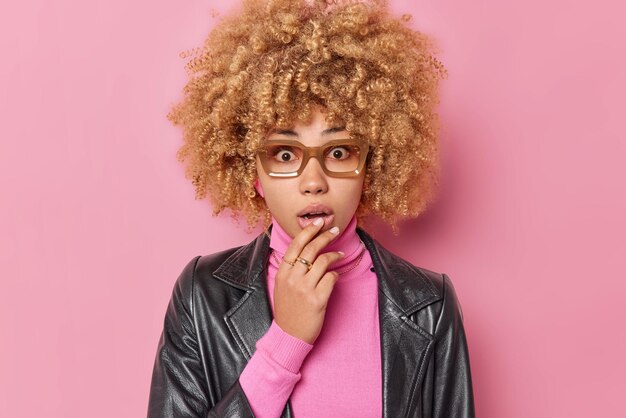 Young beautiful woman feels scared keeps handon chin stares shocked at camera hears amazing news wears spectacles turtleneck and leather jacket isolated over pink background Negative emotions