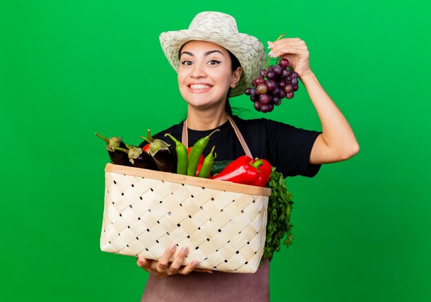 Молодая красивая женщина-садовник в фартуке и шляпе держит корзину, полную овощей и винограда, счастливая и позитивная, стоя над зеленой стеной