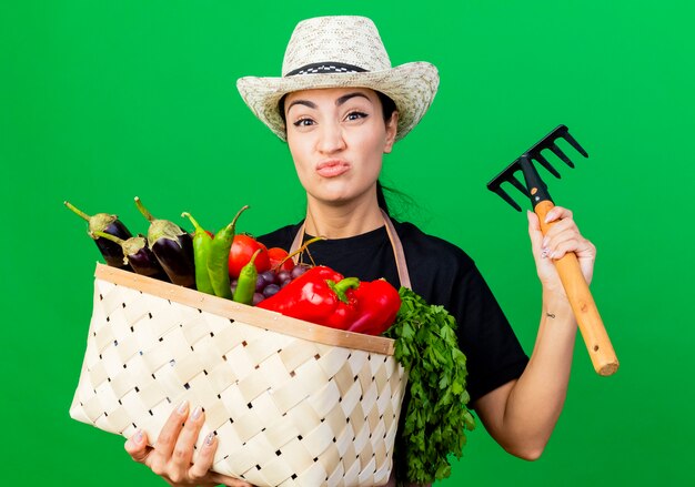 Young beautiful woman gardener in apron and hat holding basket full of vegetables and mini rake looking at front being displeased standing over green wall