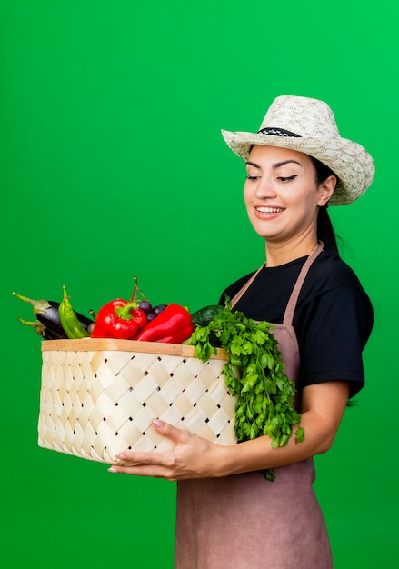 Young beautiful woman gardener in apron and hat holding basket full of vegetables smiling with happy face standing over green wall