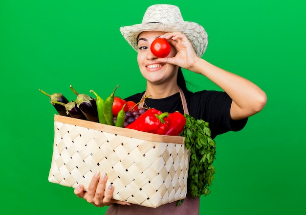 Young beautiful woman gardener in apron and hat holding basket full of vegetables and tomato near her eye smiling standing over green wall