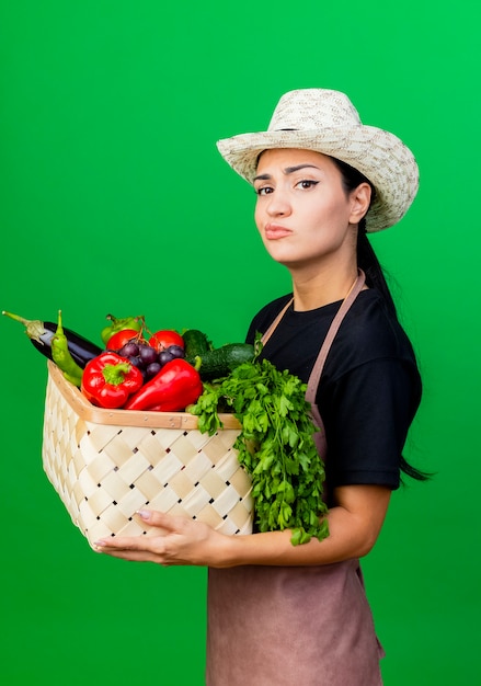 懐疑的な表現と野菜でいっぱいのバスケットを保持しているエプロンと帽子の若い美しい女性の庭師