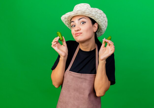 Young beautiful woman gardener in apron and hat holding broken green chili pepper with skeptic expression 