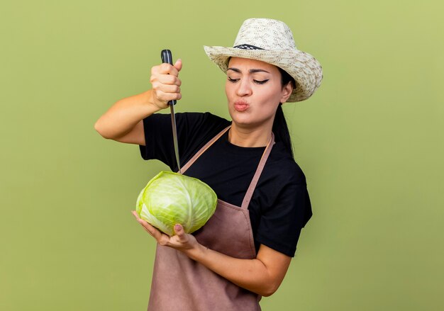 Young beautiful woman gardener in apron and hat holding cabbage and kitchen knife looking confused standing over light green wall