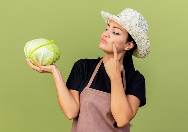 Young beautiful woman gardener in apron and hat holding cabbage looking at it with pensive expression thinking standing over light green wall