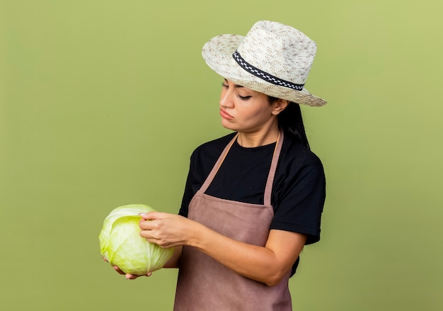 Young beautiful woman gardener in apron and hat holding cabbage looking at it with serious face standing over light green wall