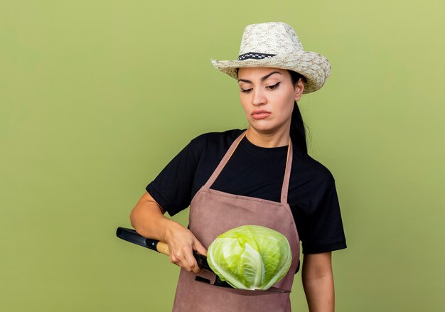 Young beautiful woman gardener in apron and hat holding cabbage on a shovel looking at it with serious face standing over light green wall