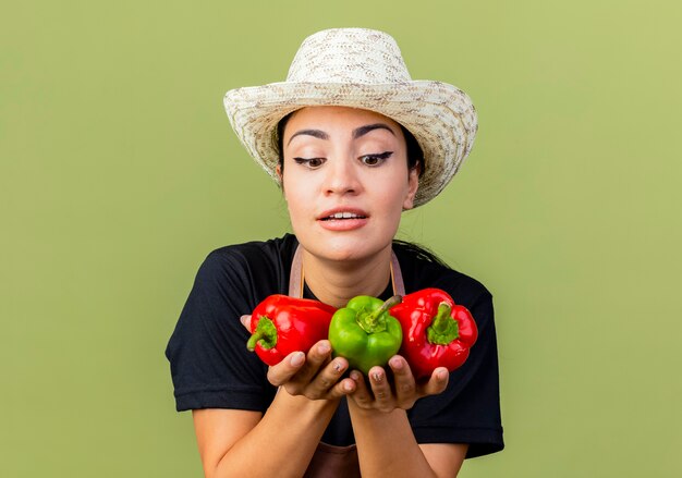 Foto gratuita giardiniere della giovane bella donna in grembiule e cappello che tengono i peperoni dolci variopinti che sorridono allegramente in piedi sopra la parete verde chiaro