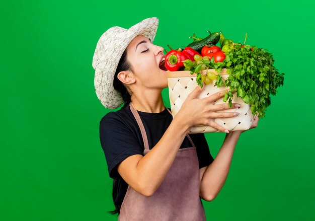 赤ピーマンを噛む野菜でいっぱいの木枠を保持しているエプロンと帽子の若い美しい女性の庭師