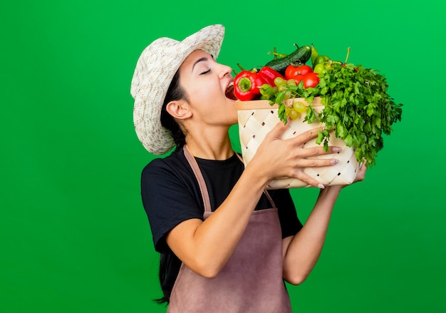 赤ピーマンを噛む野菜でいっぱいの木枠を保持しているエプロンと帽子の若い美しい女性の庭師