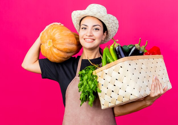 Young beautiful woman gardener in apron and hat holding crate full of vegetables and pumpkin lookign at camera with happy face 