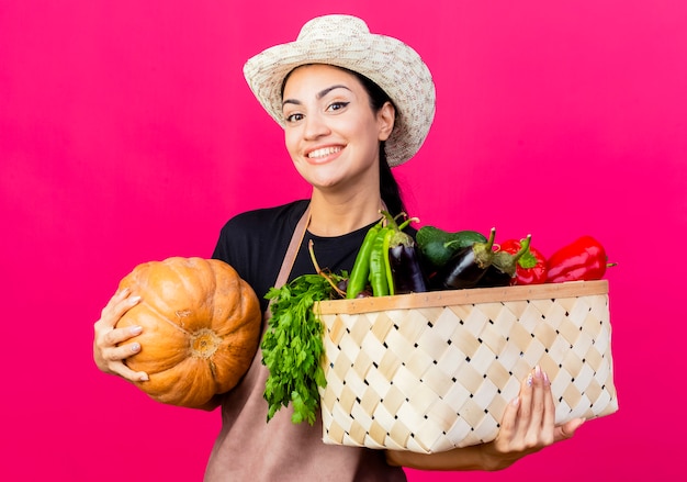 エプロンと帽子の若い美しい女性の庭師は、幸せそうな顔で笑顔の野菜とカボチャでいっぱいの木枠を保持しています