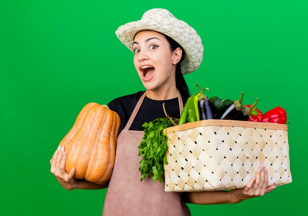 Молодая красивая женщина-садовник в фартуке и шляпе, держащая ящик, полный овощей и тыквы, счастлива и взволнована