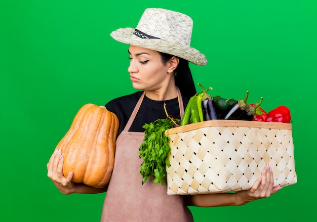 Молодая красивая женщина-садовник в фартуке и шляпе держит ящик, полный овощей и тыквы, смотрит на него с серьезным лицом