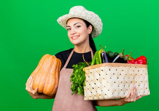 Young beautiful woman gardener in apron and hat holding crate full of vgetables and pumpkin smiling with happy face 