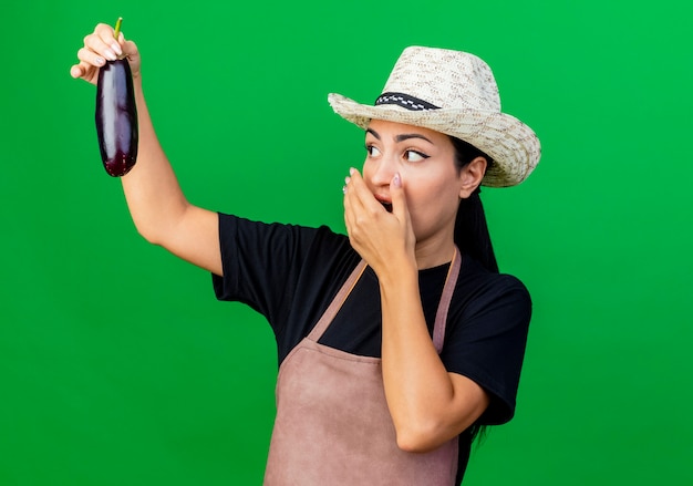 Young beautiful woman gardener in apron and hat holding eggplant looking at it being surprised and amazed standing over green wall