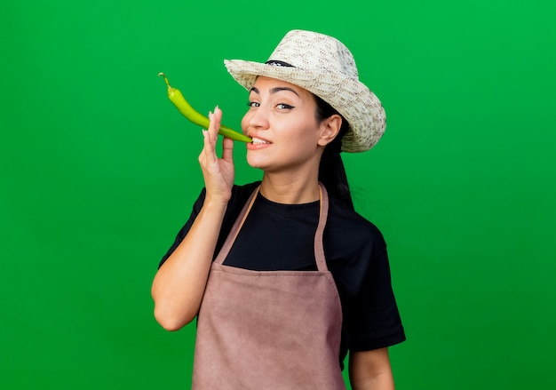 Free photo young beautiful woman gardener in apron and hat holding green chili pepper like a cigarette smiling standing over green wall