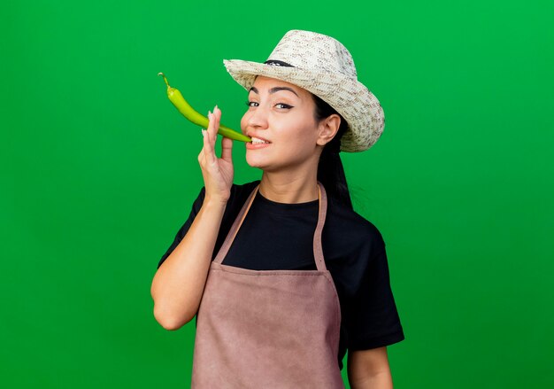 Young beautiful woman gardener in apron and hat holding green chili pepper like a cigarette smiling standing over green wall