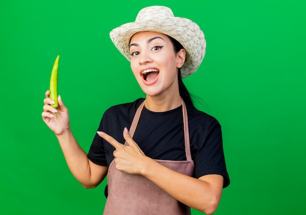 Young beautiful woman gardener in apron and hat holding green chili pepper pointign with finger at it smiling standing over green wall