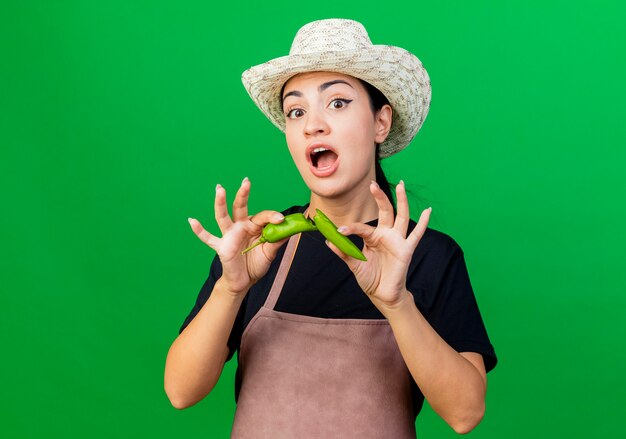 Young beautiful woman gardener in apron and hat holding green chili pepper smiling surprised 