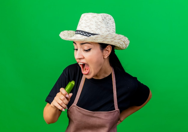 Giardiniere della giovane bella donna in grembiule e cappello che tengono peperoncino verde caldo che prova a morderlo che sta sopra la parete verde