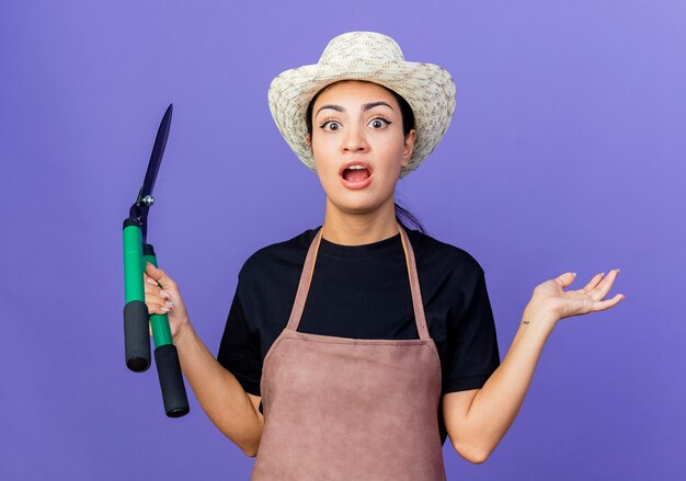 Young beautiful woman gardener in apron and hat holding hedge clippers looking surprised and amazed standing over blue wall