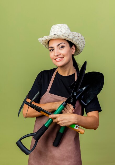 Молодая красивая женщина-садовник в фартуке и шляпе держит ножницы для живой изгороди, мотыгу и лопату, улыбаясь счастливым лицом, стоящим над светло-зеленой стеной