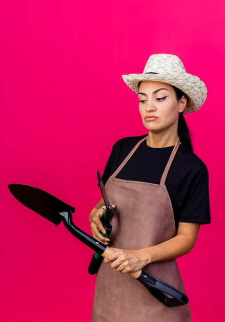 Молодая красивая женщина-садовник в фартуке и шляпе, держащая ножницы для живой изгороди и лопату, смотрит в сторону с серьезным лицом, стоящим над розовой стеной