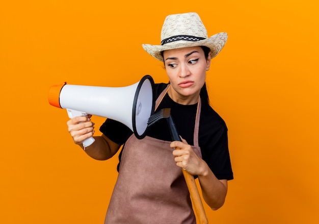 Giardiniere della giovane bella donna in grembiule e cappello che tiene mini rastrello e megafono che sembra confuso in piedi sopra la parete arancione