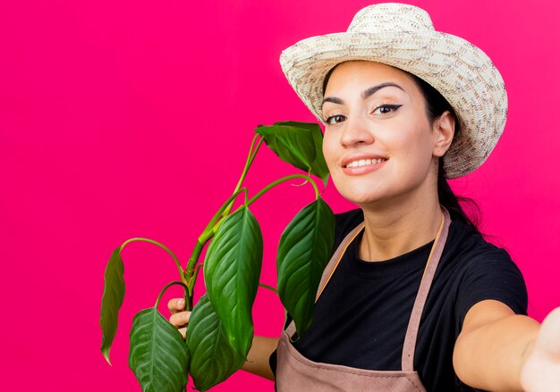 Giardiniere della giovane bella donna in grembiule e cappello che tiene pianta guardando la parte anteriore sorridente con la faccia felice in piedi sopra la parete rosa