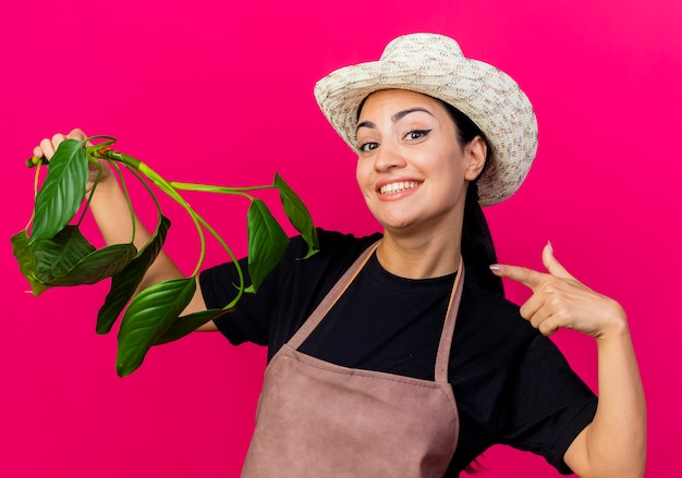 エプロンと帽子をかぶった若い美しい女性の庭師は、ピンクの壁の上に立って微笑んで人差し指で植物を指します