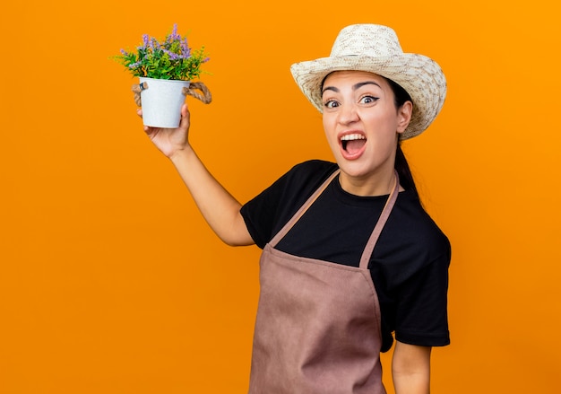 Young beautiful woman gardener in apron and hat holding potted plant looking at front being confused and displeased standing over orange wall