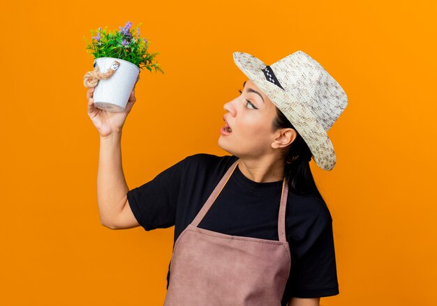 Young beautiful woman gardener in apron and hat holding potted plant looking at it being surprised and amazed standing over orange wall