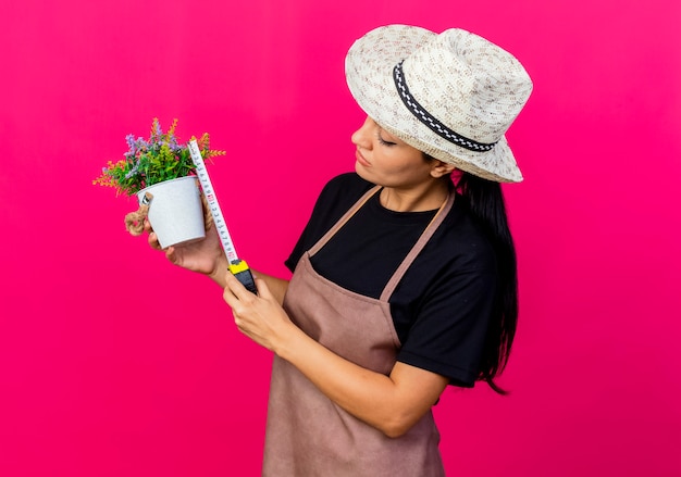 Young beautiful woman gardener in apron and hat holding potted plant and measure tape looking at it with serious face 