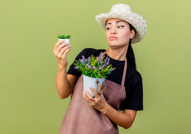 薄緑色の壁の上に立って選択をしようとして混乱しているように見える鉢植えの植物を保持しているエプロンと帽子の若い美しい女性の庭師