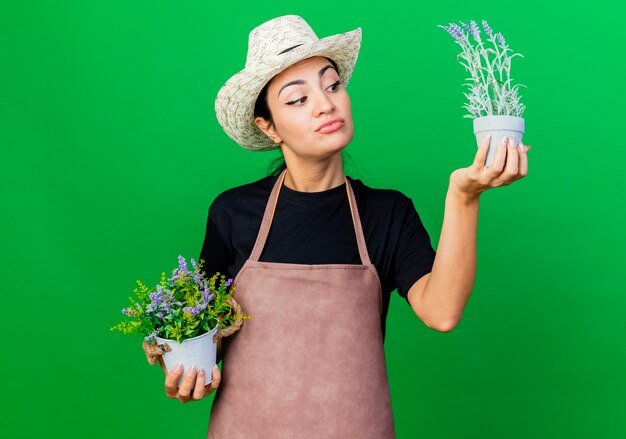 Young beautiful woman gardener in apron and hat holding potted plants looking at them being confused 