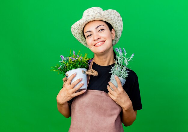 エプロンと帽子の若い美しい女性の庭師は幸せそうな顔で笑って鉢植えの植物を保持しています