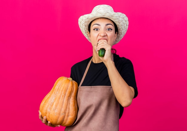 Free photo young beautiful woman gardener in apron and hat holding pumpkin and cucumber biting it standing over pink wall