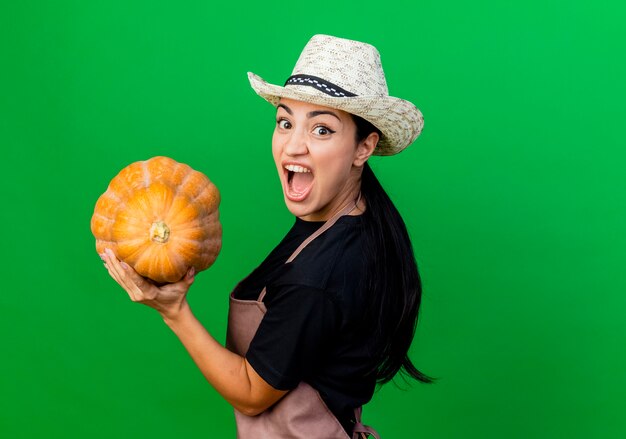 Young beautiful woman gardener in apron and hat holding pumpkin looking at front emotional and excited standing over green wall