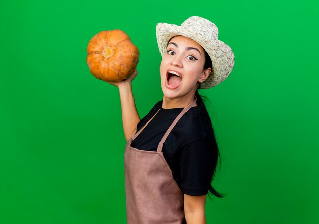 Young beautiful woman gardener in apron and hat holding pumpkin looking at front happy and excited standing over green wall