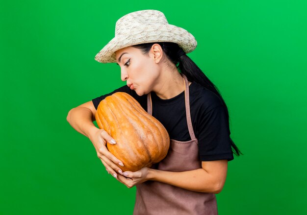 Young beautiful woman gardener in apron and hat holding pumpkin looking at it smiling and kissing it 