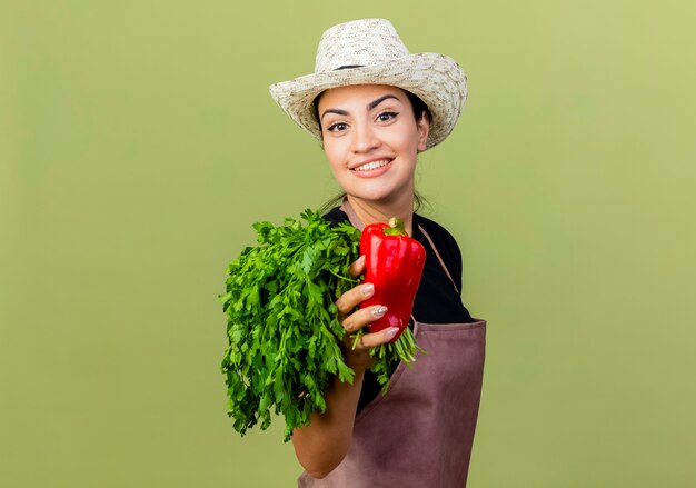 Молодая красивая женщина-садовник в фартуке и шляпе держит красный болгарский перец и свежие травы, весело улыбаясь, стоя над светло-зеленой стеной