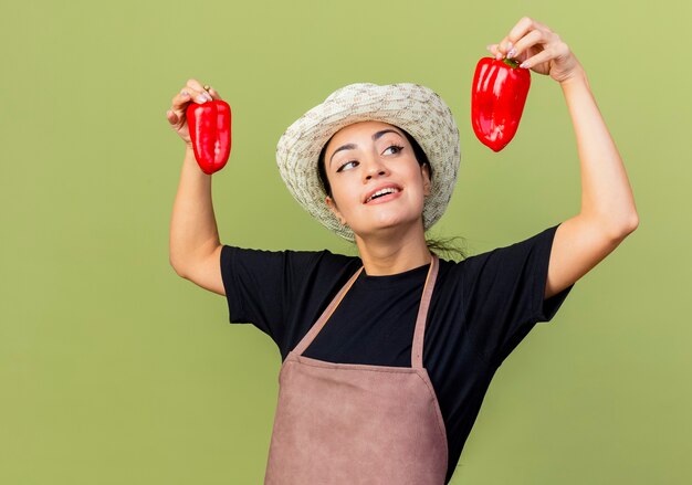 Young beautiful woman gardener in apron and hat holding red bell peppers smiling cheerfully standing over light green wall