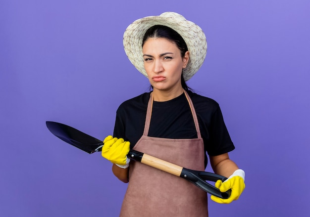 Young beautiful woman gardener in apron and hat holding shovel looking at front with frowning face standing over blue wall