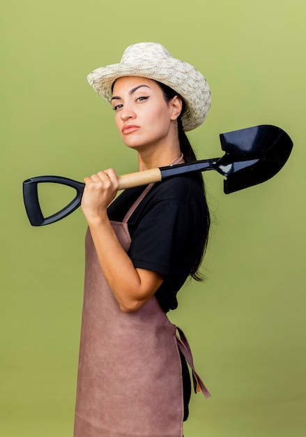 Молодая красивая женщина-садовник в фартуке и шляпе, держащая лопату, смотрит вперед с серьезным лицом, стоящим над светло-зеленой стеной