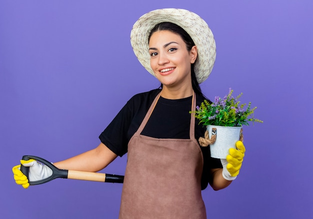 Young beautiful woman gardener in apron and hat holding shovel showing potted plant smiling cheerfully standing over blue wall