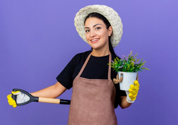 エプロンと帽子をかぶった若い美しい女性の庭師は、青い壁の上に元気に立って笑顔の鉢植えの植物を示すシャベルを保持しています