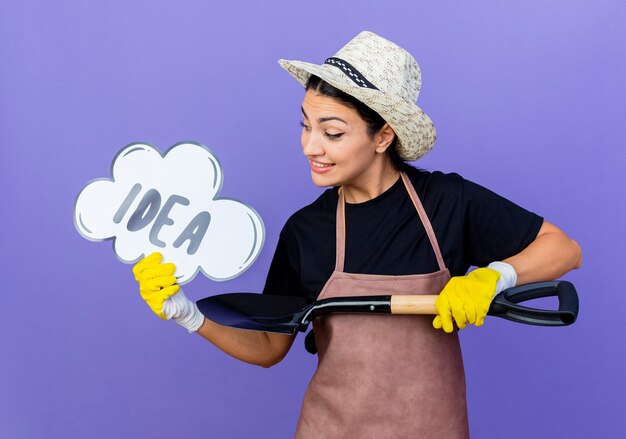 Young beautiful woman gardener in apron and hat holding shovel and speech bubble sign with word idea smiling standing over blue wall